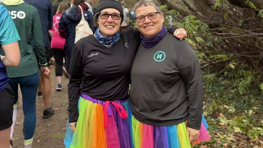 Two parkrun volunteers wearing rainbow tutus pose together on a tree-lined path, with other runners and walkers in the background.
