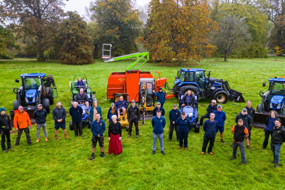 University Parks staff pose with tractors, utility vehicles and maintenance equipment on a lawn, with autumn trees behind them.